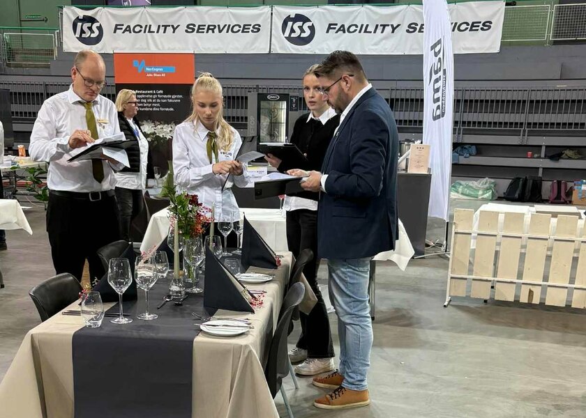 To menn og to kvinner står rundt et pent dekket bord i en messehall - Klikk for stort bilde