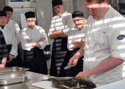 Kokk viser hvordan man fileterer kveite foran fem elever på skolekjøkken - Klikk for stort bilde