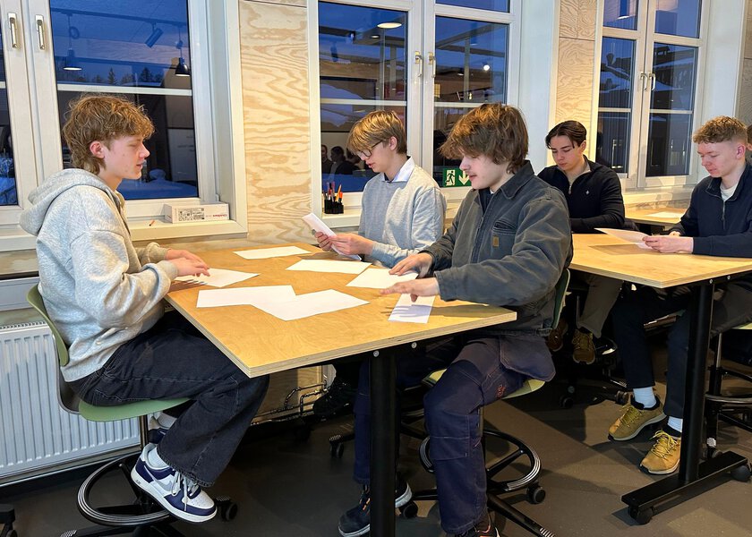 Tre elever rundt et bord og to elever rundt et bord bak - Klikk for stort bilde